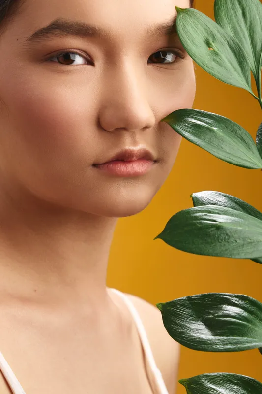 asian woman with calm expression gazes at camera, plant leaves frame her face, warm yellow backdrop, beauty portrait conveying elegance and serenity.