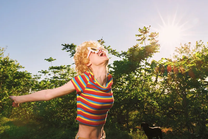 Woman lifestyle outdoors with retro film color, blonde hair, striped top, smiling under bright sunlight, enjoying nature, cheerful mood, vibrant warmth, candid park portrait