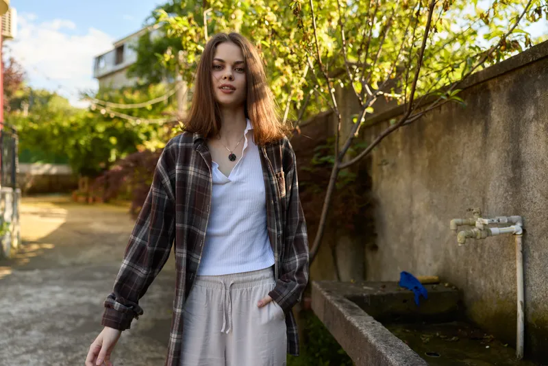Young woman with brown hair walks in a sunlit garden wearing a plaid shirt over a white tee and gray sweatpants, exuding relaxed casual style
