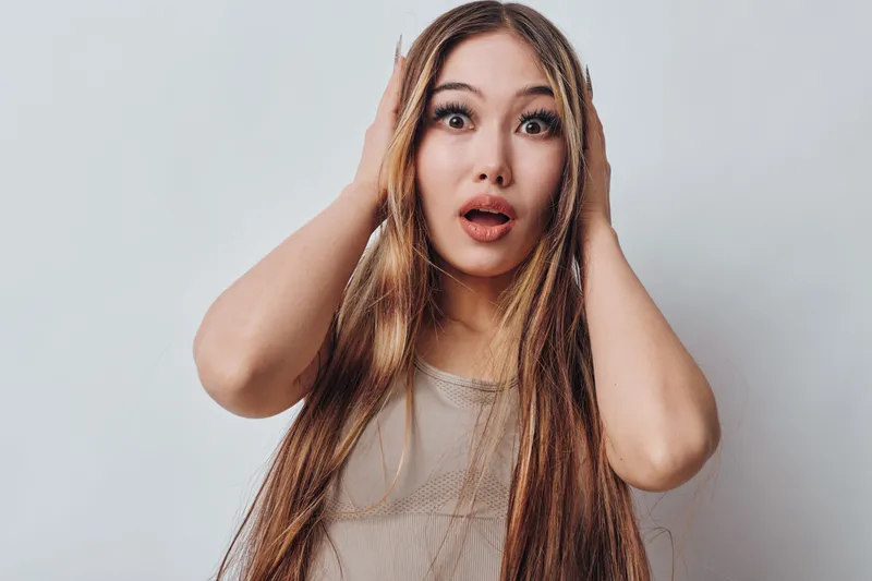 Young woman with long braided hair looks surprised and shocked with hands on head against plain background. Portrait of female with wide open eyes and mouth expression.