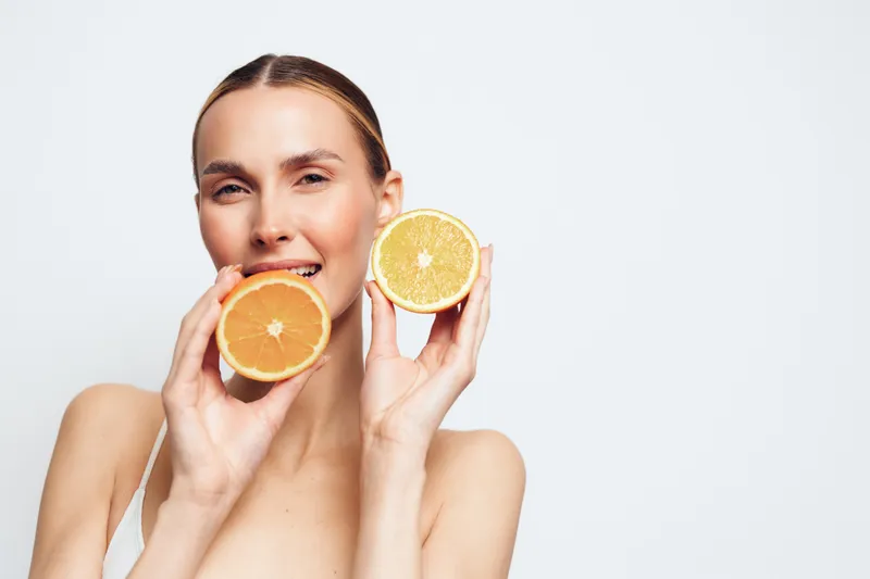 Smiling woman holding two halves of a fresh orange near her face. Close-up portrait on a white background. Concept of healthy lifestyle, freshness, and natural beauty.