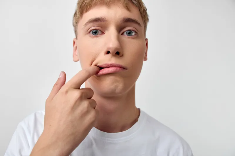Thinking young man with finger on lip, puzzled expression, casual white tee, light background, candid moment of doubt, curiosity and humor, everyday portrait style without branding