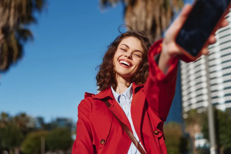 Smiling woman takes a selfie outdoors in a sunny urban setting, wearing a red coat, capturing cheerful mood and modern street style on a bright day