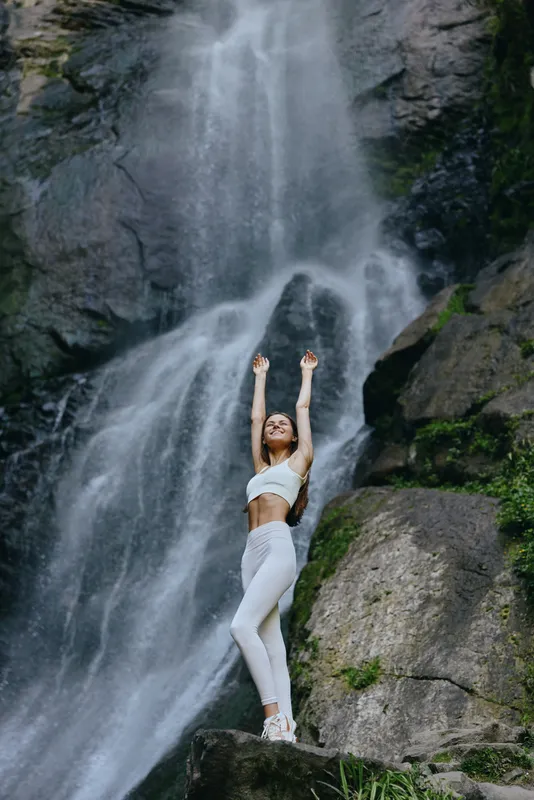 young woman stretching near waterfall in activewear, enjoying nature, wellness, and outdoor adventure, lush green rocks surrounding, serene mood.