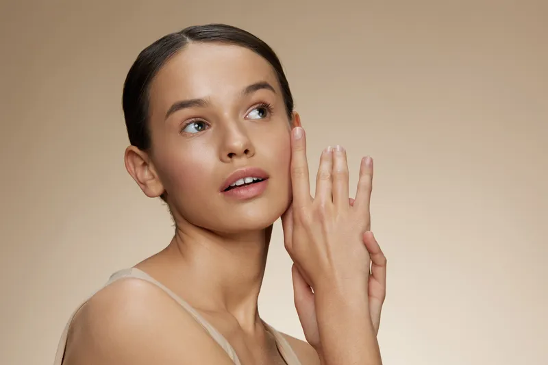 Young woman with flawless skin gently touching her face, expressing serenity and confidence against a soft beige background, ideal for skincare concepts