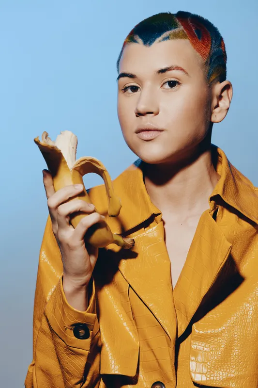 young woman with colorful shaved hair holding peeled banana, wearing yellow jacket against blue background, creative fashion portrait with vibrant style