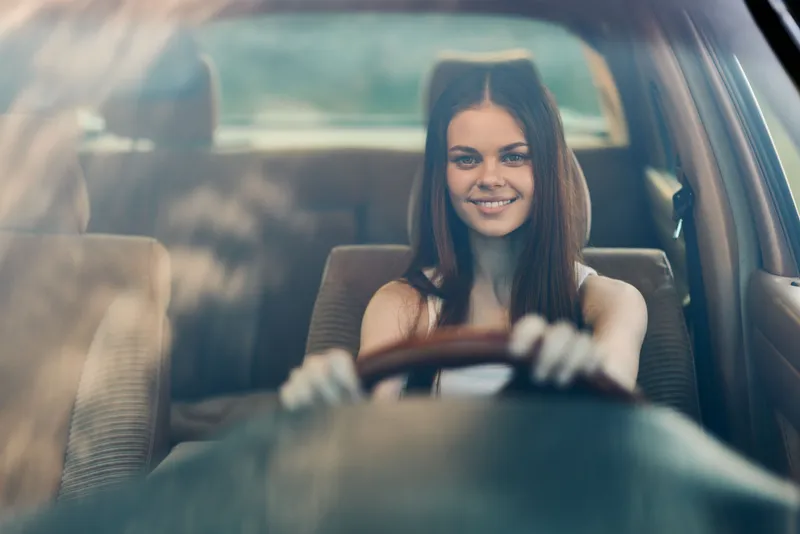 Young woman driving a car with a joyful smile, showcasing confidence in a bright interior, perfect for themes of freedom and adventure.