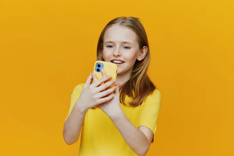 Happy young girl smiling while taking a selfie with a smartphone against a bright yellow background, wearing a cheerful yellow shirt, showcasing joy and youthfulness