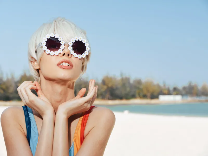 A stylish woman wearing oversized daisy sunglasses poses on a sunny beach, highlighting bold summer fashion, carefree attitude, and vibrant light with a relaxed pose.