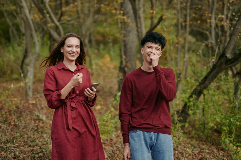 Authentic moment of two friends outdoors in an autumn forest, smiling naturally as one holds a phone and the other rests a hand near the face, candid and credible.