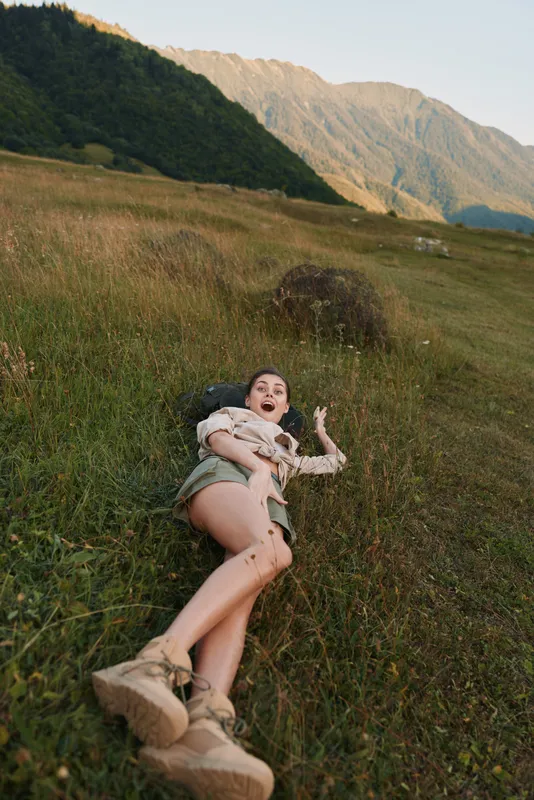 Young woman lying in a grassy field with a mountain backdrop, expressing carefree joy.