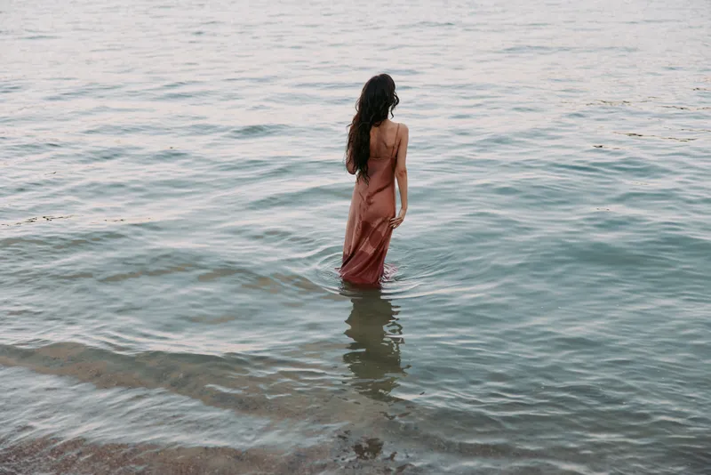 A lone figure stands waist deep in calm water, wearing a flowing dress, with a warm horizon glow and gentle reflection, evoking calm solitude.