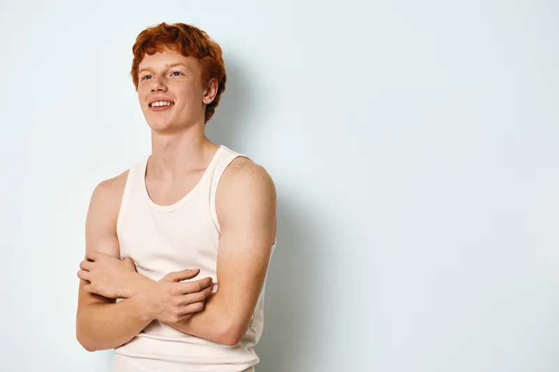 Young man with red hair wearing white tank top smiling and crossing arms on light background. Casual portrait with natural expression and relaxed body language in studio setting.