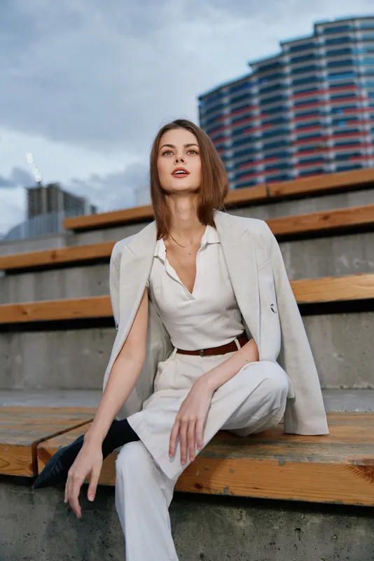 Confident woman in stylish white outfit sitting outdoors in urban environment with modern architecture and moody sky, showing elegance and calm