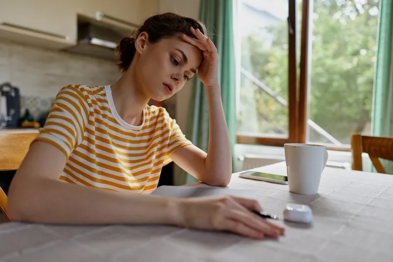 Thoughtful woman indoors, resting hand on her forehead at home desk with notebook and mug, daytime light, striped shirt, reflective mood, casual lifestyle, focused moment