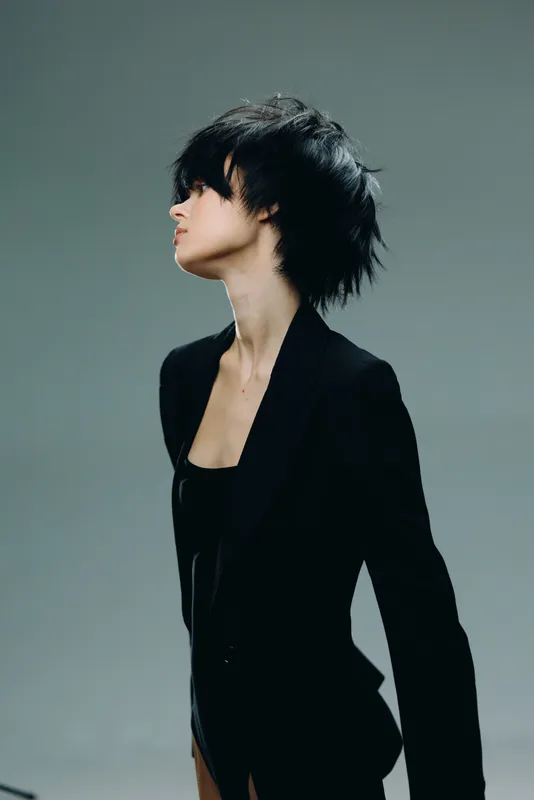 Profile of a young woman with short black hair wearing a black blazer against a solid grey background.