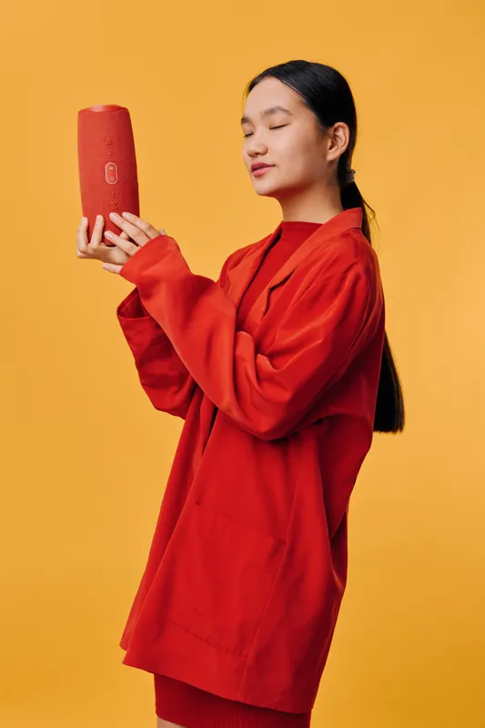 Woman holding red portable speaker with closed eyes, wearing red oversized sweater in front of orange background. Modern technology, music lover, casual style and lifestyle concept.