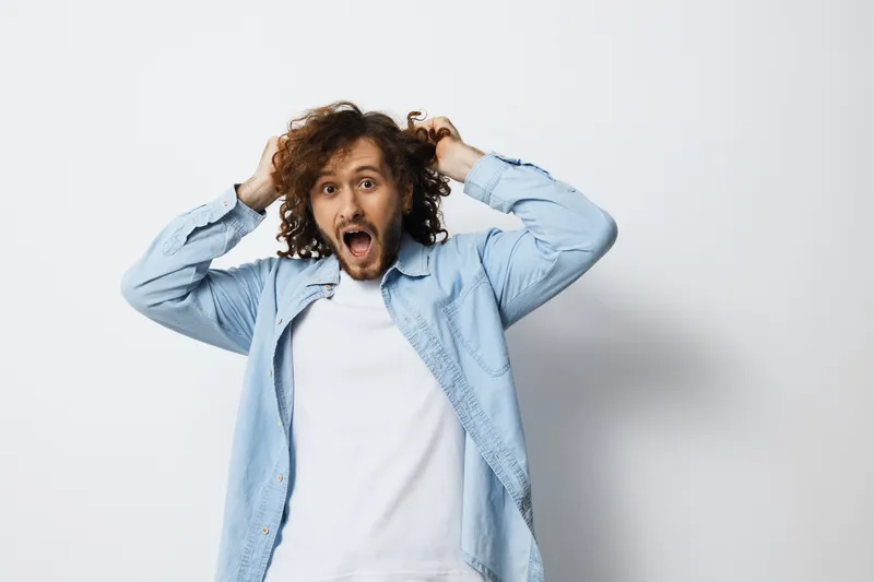Excited young man with curly hair expressing joy in a casual denim shirt against a light background, representing emotion and youthful energy