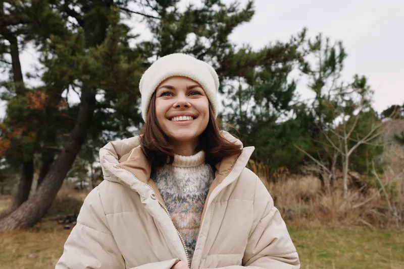 Smiling woman outdoors in winter attire, wearing a beige puffer jacket and knit beanie, standing in a natural landscape with trees. Cozy fashion and chilly weather mood for seasonal portraits.