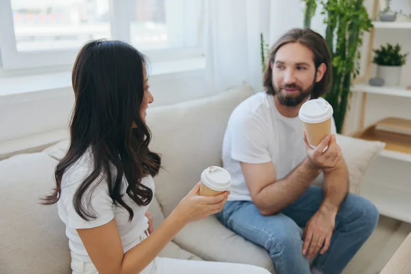 Мужчина и женщина сидят дома на диване в белых стильных футболках, пьют кофе из крабовых стаканчиков из кофейни и весело общаются, смеются и улыбаются
