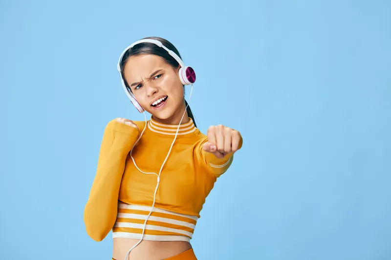 Energetic fitness woman in orange athleticwear dances with headphones against a bright blue studio backdrop, showcasing movement, confidence, and sporty motivation. Her pose conveys power and rhythm,