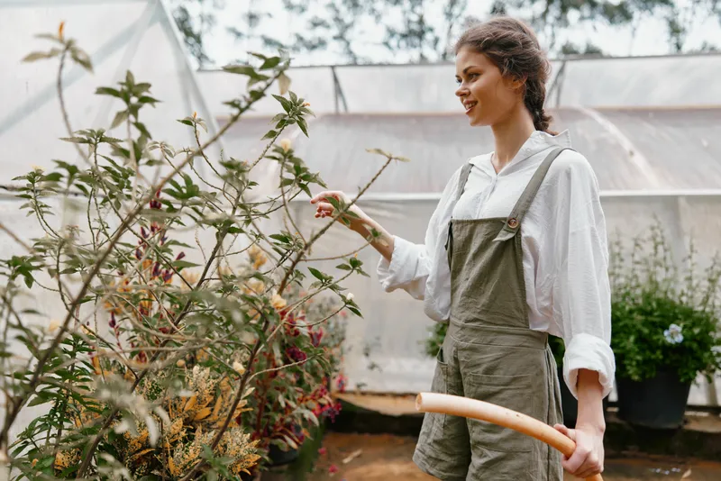Young woman watering plants in a greenhouse, wearing casual overalls and a white shirt, showcasing a love for gardening and nature