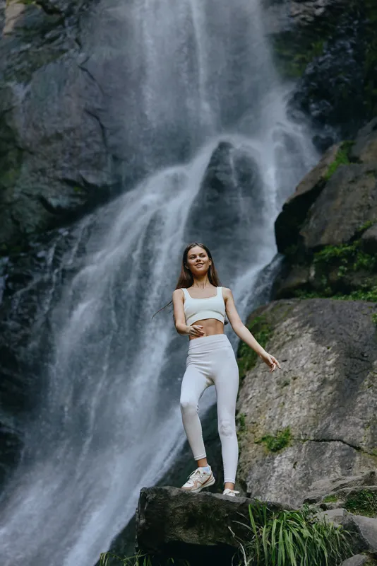 Active woman in white sportswear standing confidently on rock near waterfall surrounded by lush greenery, showcasing fitness and outdoor adventure in nature.