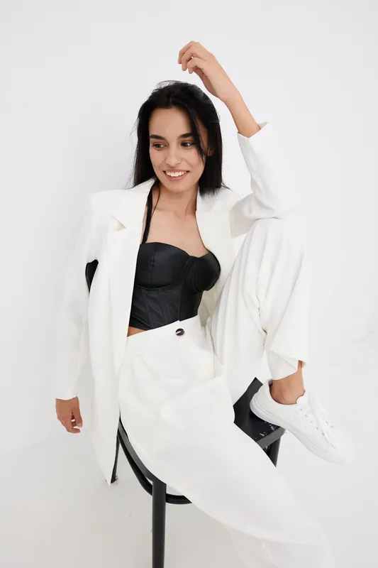 Smiling woman in white suit and black top poses on chair with relaxed confidence, delivering modern fashion style with casual sneakers against plain white background.