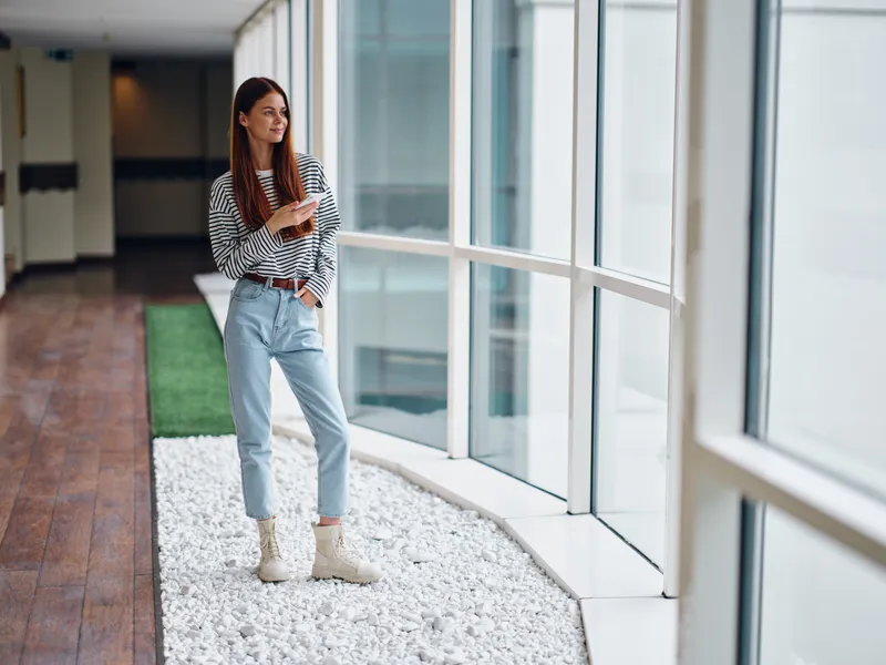 Женщина стоит у окна в полный рост и держит телефон с улыбкой, онлайн-работа через интернет, фриланс