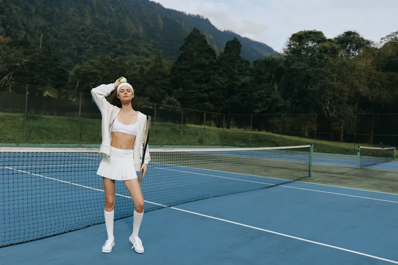 Young athletic woman in stylish white sportswear poses confidently on a tennis court surrounded by lush green mountains, showcasing a vibrant outdoor lifestyle