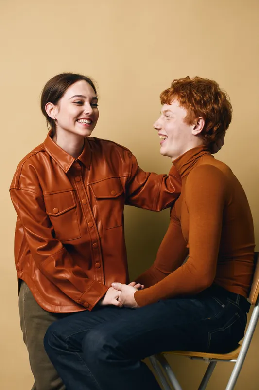 Two young people smiling and talking indoors, casual outfit, warm colors, friendship and positive emotions, sitting and standing with eye contact and happiness.