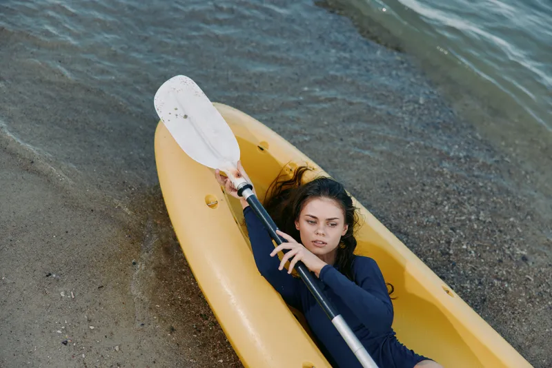A young woman sits in a yellow kayak near the shoreline, gripping a paddle as calm waves lap the sandy beach, capturing a peaceful outdoor water scene.