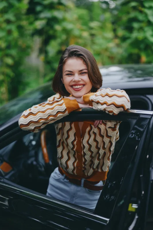 A joyful woman leans out of a car window, smiling at the camera, wearing a colorful striped knit sweater.