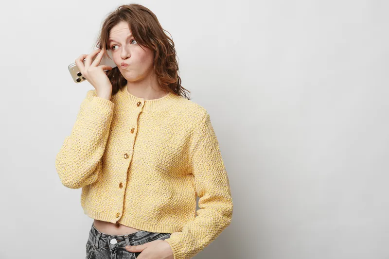 Young woman in yellow knitted sweater holds smartphone near face, making funny expression. Casual style, isolated on white background. Concept of communication, technology, and emotions.