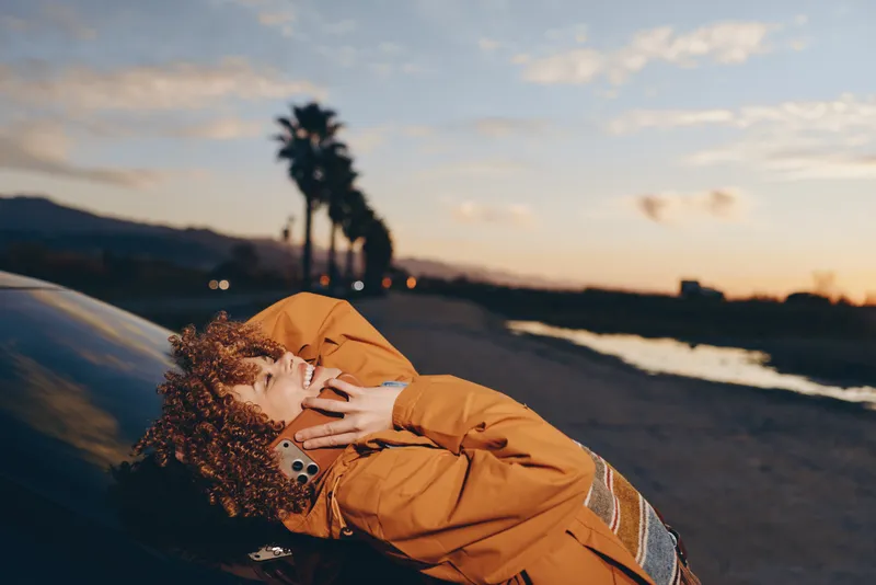 Woman lifestyle at sunset on a coastal road, hugging a curly dog in an orange jacket, golden light and warm wind show tender companionship and calm outdoors moments for pet care