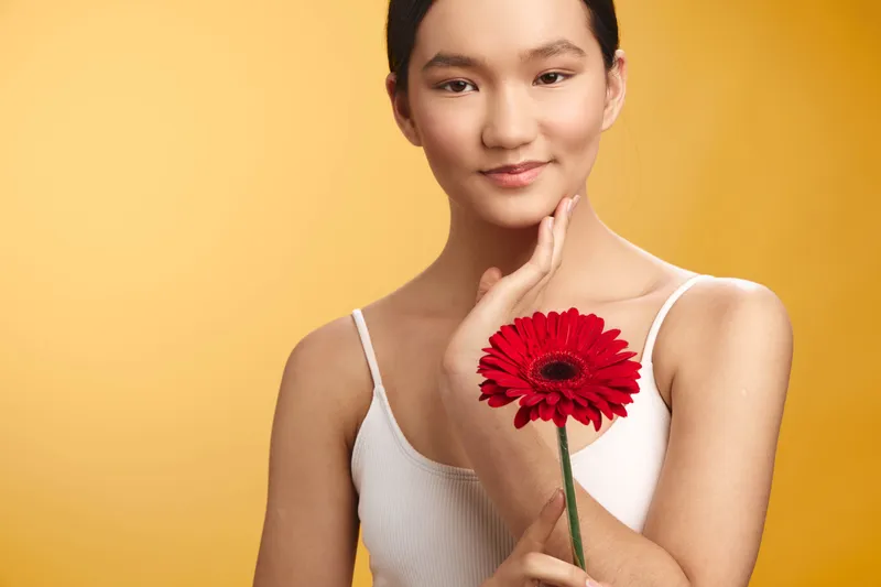 asian woman gently holds a vibrant red flower against a bright yellow backdrop, presenting a poised pose, soft smile, and serene confidence that radiates warmth and charm.