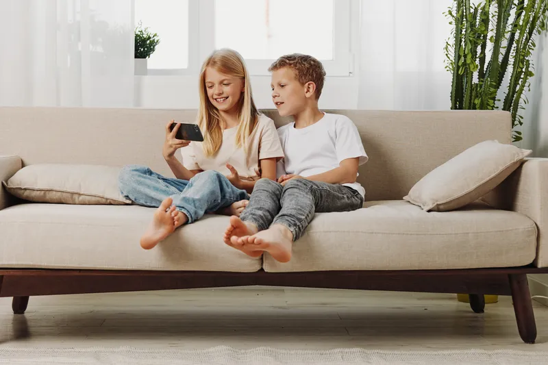 Two happy children sitting barefoot on a beige sofa, watching a smartphone together. Bright living room setting with plants and natural light. Concept of childhood, technology, and family.