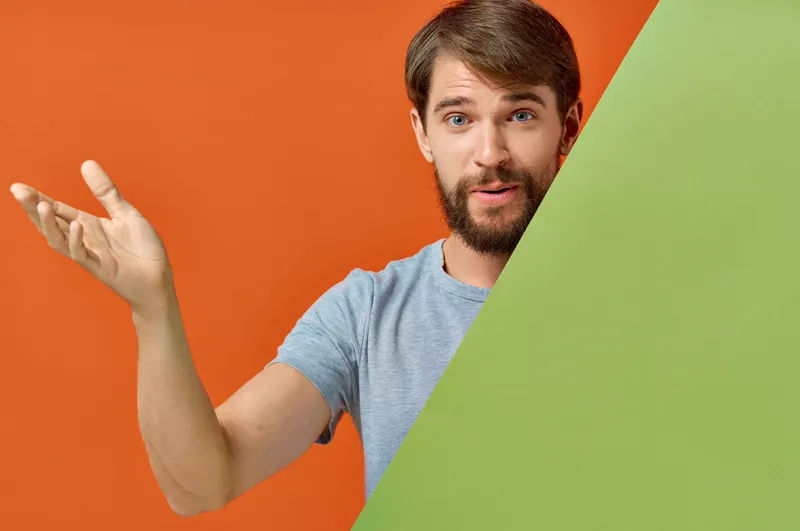 Бородатый мужчина в серой футболке держит зеленый макет плаката на оранжевом фоне