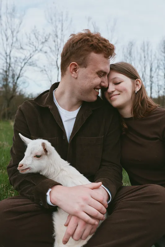 Couple outdoors with a gentle goat kid, smiling and close, warm light, relaxed pose, rustic setting, soft breeze, natural colors, candid affection, peaceful countryside moment
