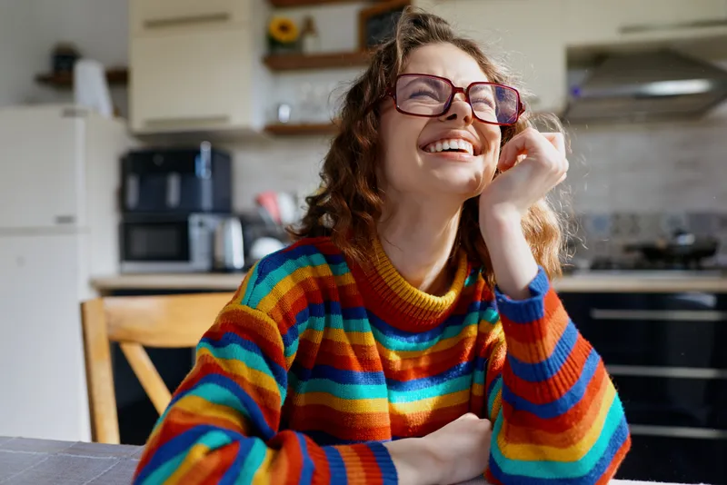 A joyful woman in a vibrant striped sweater sits at a bright kitchen table, smiling with glasses, capturing a warm, casual home lifestyle moment that feels relatable, cozy, spontaneous, creative, and
