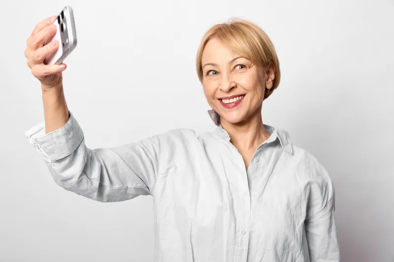 Senior woman takes selfie with smartphone, smiling portrait in clean white background studio, communication and technology for online sharing, cheerful confidence and wellbeing