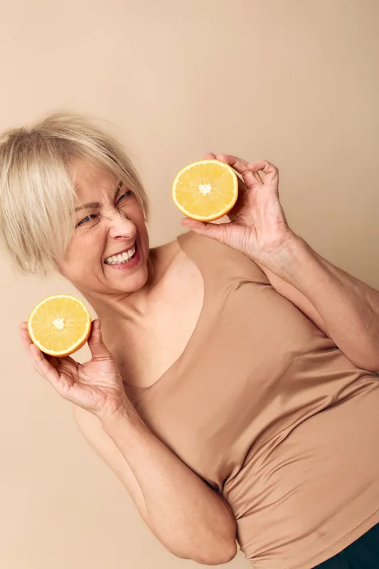Happy mature woman with short blonde hair holds two halves of fresh orange, smiling and posing on beige background. Concept of healthy lifestyle, vitamin c and natural nutrition.