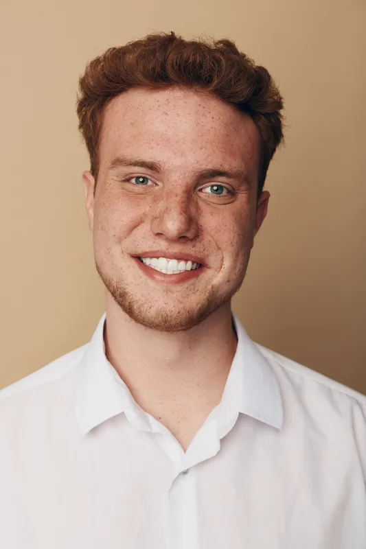 Smiling young man with red hair and freckles wearing white shirt looks at camera on beige background. Portrait of confident male model with natural skin and friendly expression.