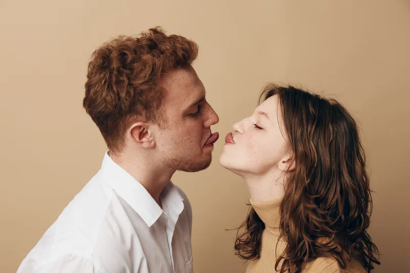 Young couple with curly hair playfully sticks out tongues facing each other on beige background. Concept of love, intimacy, fun, and relationship moments between man and woman.