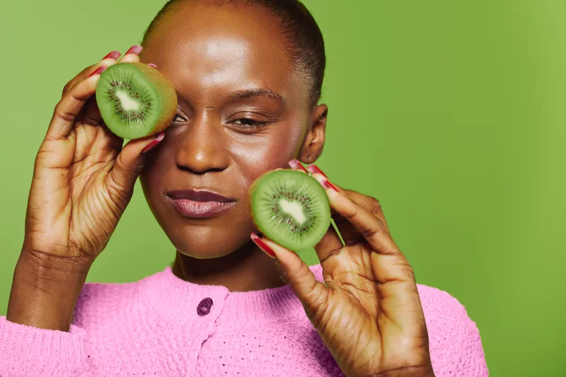 Smiling African woman with dark skin holds two halves of kiwi fruit near face on green background. Concept of healthy eating, natural food, wellness, and fresh fruit lifestyle.