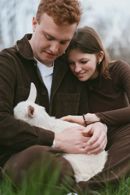 A loving couple sits outdoors with a gentle goat kid, sharing a tender moment in nature, embodying lifestyle and companionship, as soft light enhances their affectionate bond.