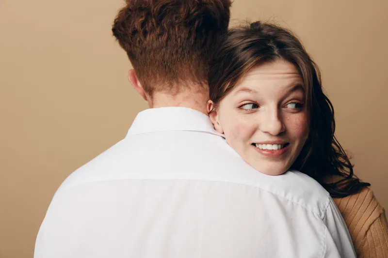 Young woman with freckles smiles happily while hugging a man in a white shirt against beige background. Concept of affection, love, and close relationship between couple.