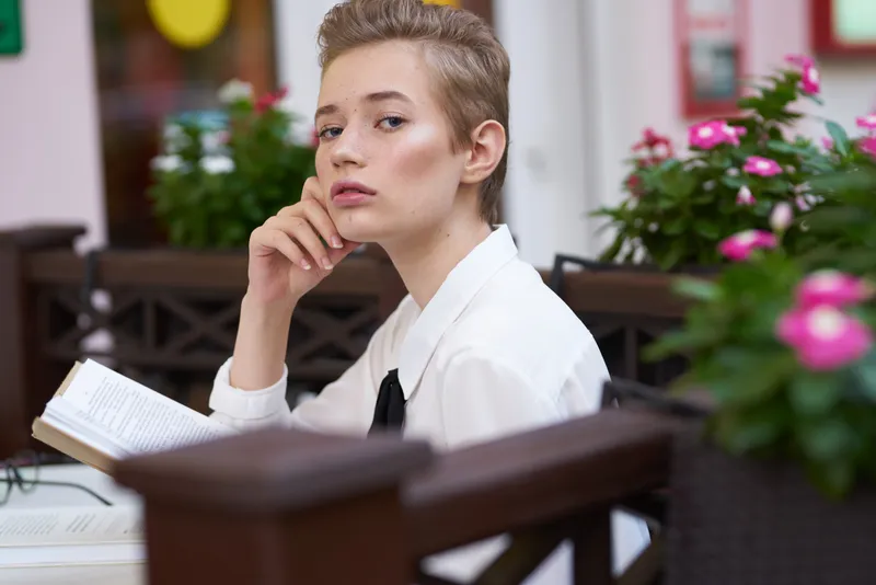 Умная женщина с книгой сидит за столиком в кафе, рядом розовые цветы, весенний макияж