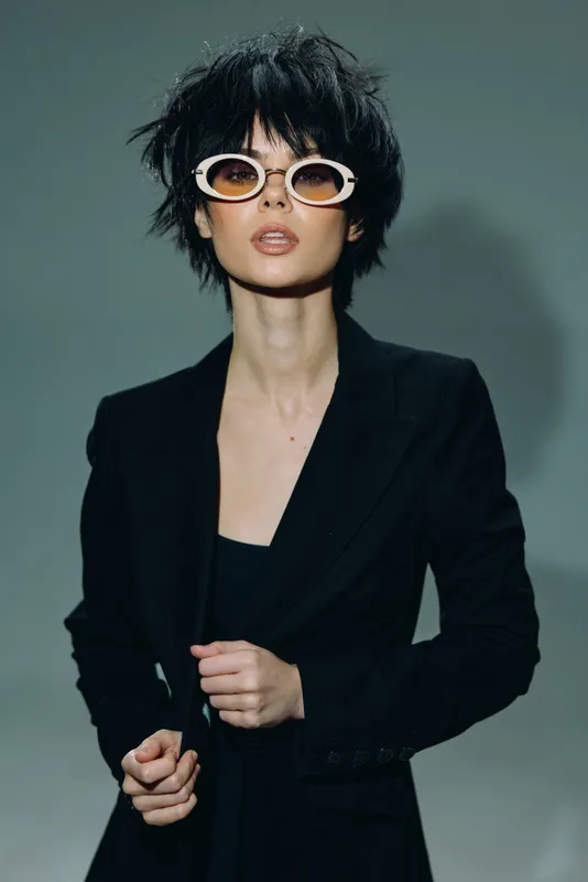 Stylish woman in black jacket wearing white sunglasses with confident expression in studio lighting
