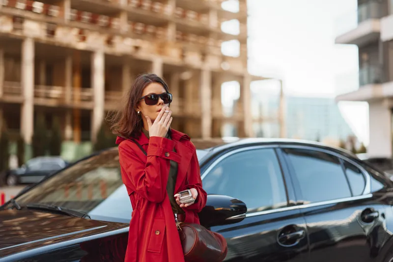 A stylish woman in a red trench coat stands beside a sleek car on a modern urban street, wearing sunglasses and exuding confidence in a chic city moment